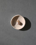 A single gold earring with a white pearl drop sits in a small beige ceramic dish on a textured gray surface, casting a strong shadow.
