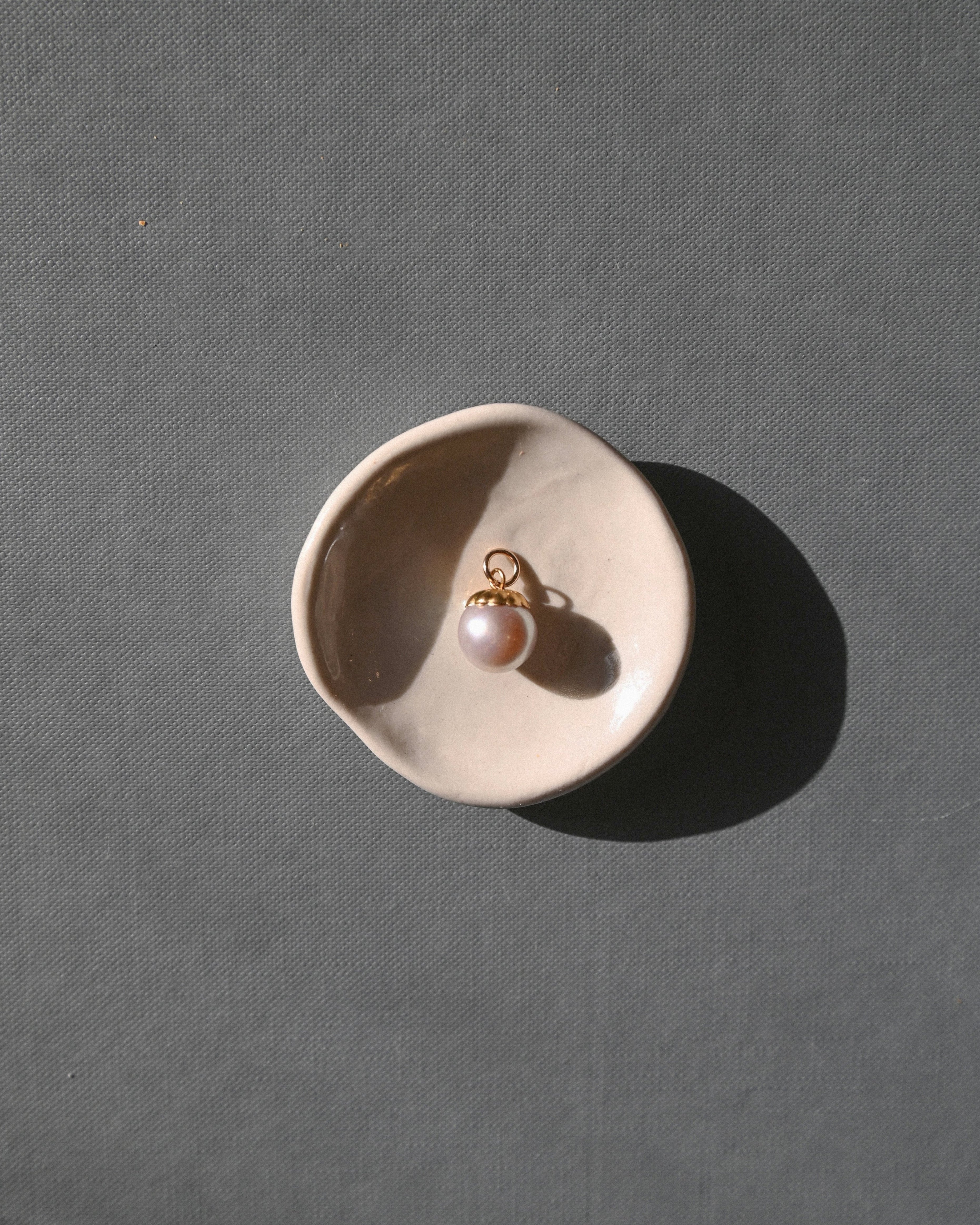 A single gold earring with a white pearl drop sits in a small beige ceramic dish on a textured gray surface, casting a strong shadow.